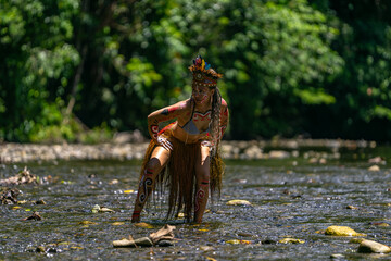 Indigenous woman crouches in a shallow river wearing tribal attire and body paint, immersed in the wild jungle
