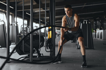 Strong and masculine man in sportswear using training ropes for exercise at gym. Athlete male working out with battle ropes in fitness center. Bodybuilding, fitness and health care concept.