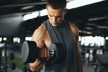 Young handsome sportsman bodybuilder weightlifter training biceps with dumbbells while looking at...
