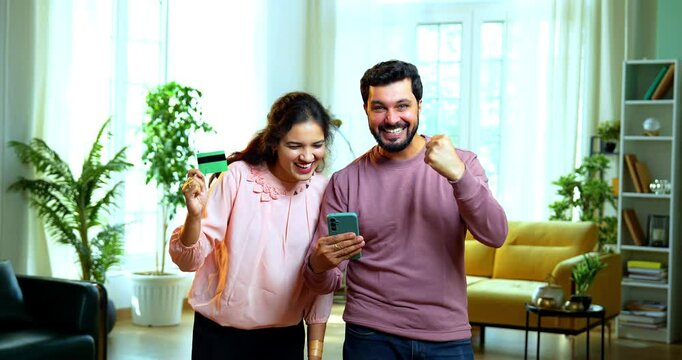 Indian Asian young couple standing in modern living room celebrating success holding credit or debit card and smartphone, with five hundred rupee Indian currency notes raining down on them