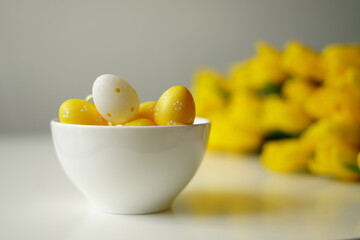 Bright Easter composition with yellow and white eggs in a white ceramic bowl next to fresh yellow tulips on a white table. Minimalist holiday still life, perfect for spring-themed content. 