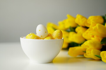 Bright Easter composition with yellow and white eggs in a white ceramic bowl next to fresh yellow tulips on a white table. Minimalist holiday still life, perfect for spring-themed content. 