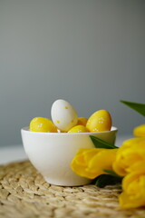 Bright Easter composition with yellow and white eggs in a white ceramic bowl next to fresh yellow tulips on a white table. Minimalist holiday still life, perfect for spring-themed content. 
