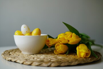 Bright Easter composition with yellow and white eggs in a white ceramic bowl next to fresh yellow tulips on a white table. Minimalist holiday still life, perfect for spring-themed content. 