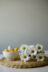  Bright and cheerful Easter-themed still life with a white ceramic bowl of yellow and white decorative eggs and fresh white daisies on a woven placemat. 