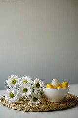  Bright and cheerful Easter-themed still life with a white ceramic bowl of yellow and white decorative eggs and fresh white daisies on a woven placemat. 