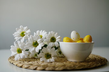  Bright and cheerful Easter-themed still life with a white ceramic bowl of yellow and white decorative eggs and fresh white daisies on a woven placemat. 