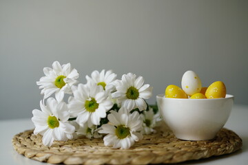  Bright and cheerful Easter-themed still life with a white ceramic bowl of yellow and white decorative eggs and fresh white daisies on a woven placemat. 