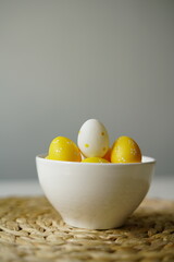 Minimal Easter-themed still life featuring yellow and white polka dot decorative eggs in a clean white ceramic bowl, placed on a woven straw placemat. perfect for illustrating Easter, spring 