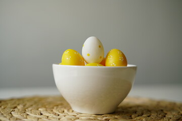 Minimal Easter-themed still life featuring yellow and white polka dot decorative eggs in a clean white ceramic bowl, placed on a woven straw placemat. perfect for illustrating Easter, spring 