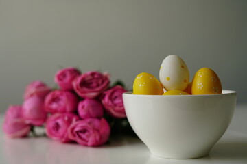 Minimalist spring composition with yellow polka dot Easter eggs in a white ceramic bowl next to a bouquet of pink roses. Clean background and soft light make this image perfect for Easter 