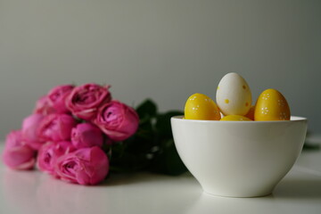 Minimalist spring composition with yellow polka dot Easter eggs in a white ceramic bowl next to a bouquet of pink roses. Clean background and soft light make this image perfect for Easter 