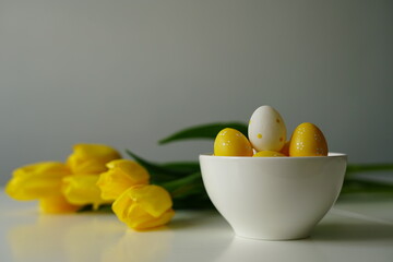 Bright Easter composition with yellow and white eggs in a white ceramic bowl next to fresh yellow tulips on a white table. Minimalist holiday still life, perfect for spring-themed content. 