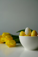 Bright Easter composition with yellow and white eggs in a white ceramic bowl next to fresh yellow tulips on a white table. Minimalist holiday still life, perfect for spring-themed content. 