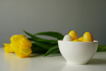 Bright Easter composition with yellow and white eggs in a white ceramic bowl next to fresh yellow tulips on a white table. Minimalist holiday still life, perfect for spring-themed content. 