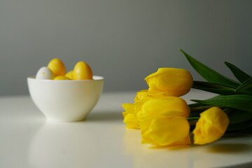 Bright Easter composition with yellow and white eggs in a white ceramic bowl next to fresh yellow tulips on a white table. Minimalist holiday still life, perfect for spring-themed content. 