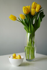 vertical photo Bright Easter composition with yellow and white eggs in a white ceramic bowl next to fresh yellow tulips on a white table. Minimalist holiday still life, perfect for spring
