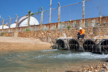 An environmental engineer in safety gear sits on a concrete drainage outlet near a factory,...