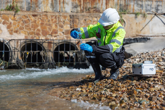A safety-clad environmental worker collects a water sample from a rocky shoreline near an industrial drainage outlet, using scientific equipment to test for pollutants or contamination.