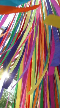 Multicolored satin ribbons flutter in wind and sunbeams sparkle through gaps. Vertical video of colorful decorative installation made of densely suspended textile strips.