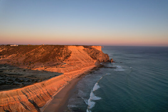 Aerial view of beautiful beach with rugged cliffs and tranquil ocean waves at sunset, Algave, Portugal.