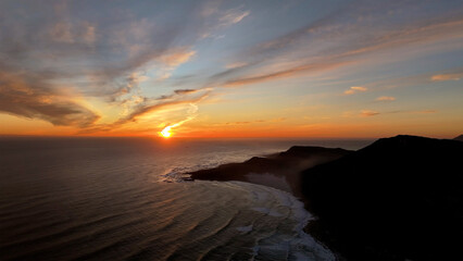 Aerial view of sunset over serene waves and majestic mountains along the coastal shoreline, Scarborough, Western Cape, South Africa.