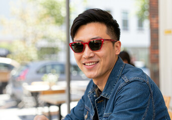 Obraz premium Portrait of a smiling asian man wearing red sunglasses while sitting at a table of an outdoor cafe