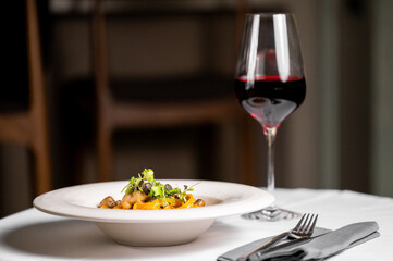 A beautifully plated pasta dish garnished with herbs sits on a table alongside a glass of red wine, creating an elegant dining experience in a cozy restaurant setting.
