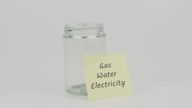 Gas, Water, Electricity text on a yellow note stuck to a jar of coins, with a hand inserting a coin. The image symbolizes saving for utility bills, including gas, water, and electricity