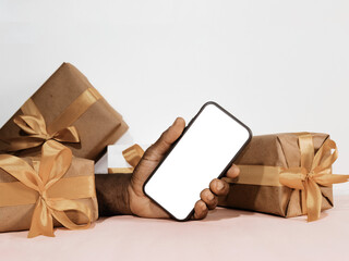 Black hand holding showing blank phone screen in his hand with gift boxes around. Mobile telephone mockup template	