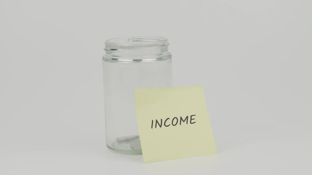 Income text on a yellow note stuck to a jar of coins, with a hand inserting a coin. The image symbolizes the act of saving and managing income for financial stability and future growth