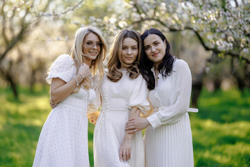 Fototapeta premium Three women pose elegantly in white dresses for a park photo