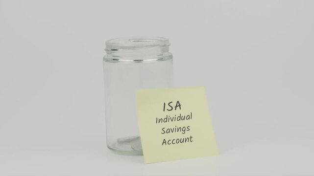 ISA (Individual Savings Account) text on a yellow note stuck to a jar of coins, with a hand inserting a coin. The image represents saving in an ISA to grow wealth and take advantage of tax benefits