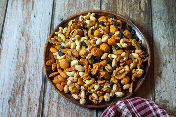 A round wooden plate holds cashews, almonds, pistachios, dried apricots, raisins, and walnuts, neatly arranged on a rustic wooden table beside a red checkered cloth.