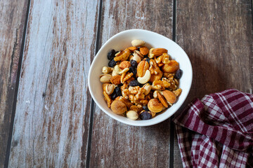 A round wooden plate holds cashews, almonds, pistachios, dried apricots, raisins, and walnuts, neatly arranged on a rustic wooden table beside a red checkered cloth.