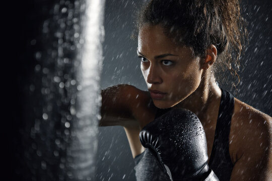 A focused woman in boxing gloves strikes a heavy bag, showcasing her strength and determination while training indoors with a rainy backdrop