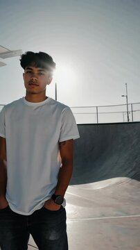 Young man poses confidently in a white T-shirt, against a skatepark bowl backdrop, enjoying the sunlight and warm weather.