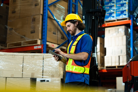 Caucasian adult male warehouse worker wearing yellow helmet using digital tablet near packages checking data during logistics operation in warehouse showing digital workflow in storage.