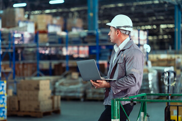 Caucasian adult male warehouse supervisor wearing white safety helmet and gray uniform using laptop...