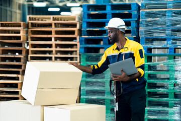 African adult male warehouse worker in safety uniform inspecting cardboard boxes while holding laptop during operation process in warehouse showing monitoring and coordination duty.