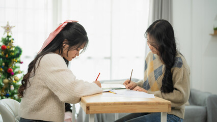Two Young Women Engaged in Art Activities at Home with Christmas Tree Decor in Background, Creating Artwork on Table with Colorful Pencils and Paints