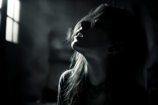 Retrato en blanco y negro de una mujer mirando hacia arriba, con luz tenue que ilumina su rostro y cabello. Ambiente melanc&oacute;lico y misterioso.