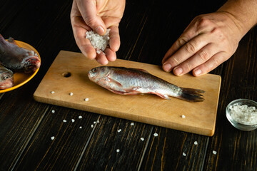 Hands season a freshly caught fish with coarse salt on a wooden cutting board. Nearby, another fish and a bowl of salt add to the kitchen atmosphere, highlighting the pre-cooking process
