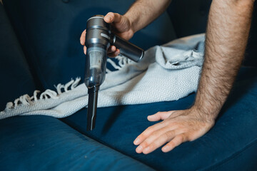Handheld vacuuming soft upholstery on blue sofa by adult male