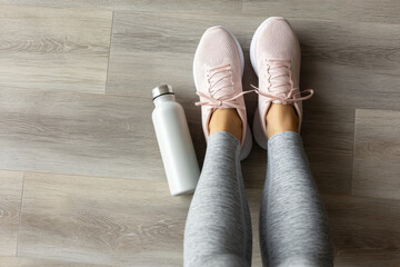 A woman wearing grey leggings and pink sneakers stands on a wooden floor next to a white water bottle, ready for a workout routine