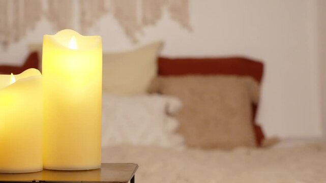 Right-to-left panning of 2 artificial candles on a golden base, in the background a futon with cushions and macrame decoration is visible in the background