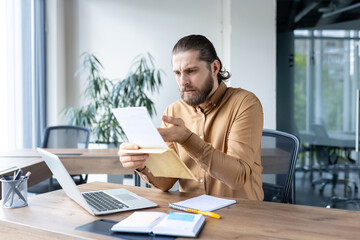 A focused businessman is looking at documents in his hands, possibly analyzing data at his desk...