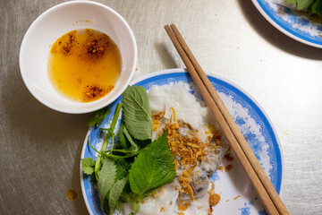 A portion of banh cuon with dipping sauce, greens, and fried shallots with chopsticks sits on a...