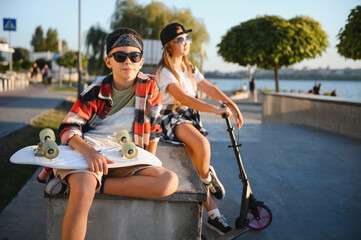 Satisfied children in the skate park. A boy with a skate and a girl with a scooter spend time together. Summer vacation