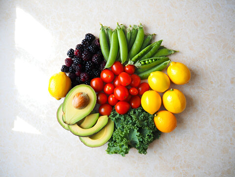 Top-down view of various fruits and vegetables laid out in a radial, circular design. Bright, colorful vegan ingredients like carrots, bell peppers, spinach, kiwi, strawberries, and broccoli. Clean st